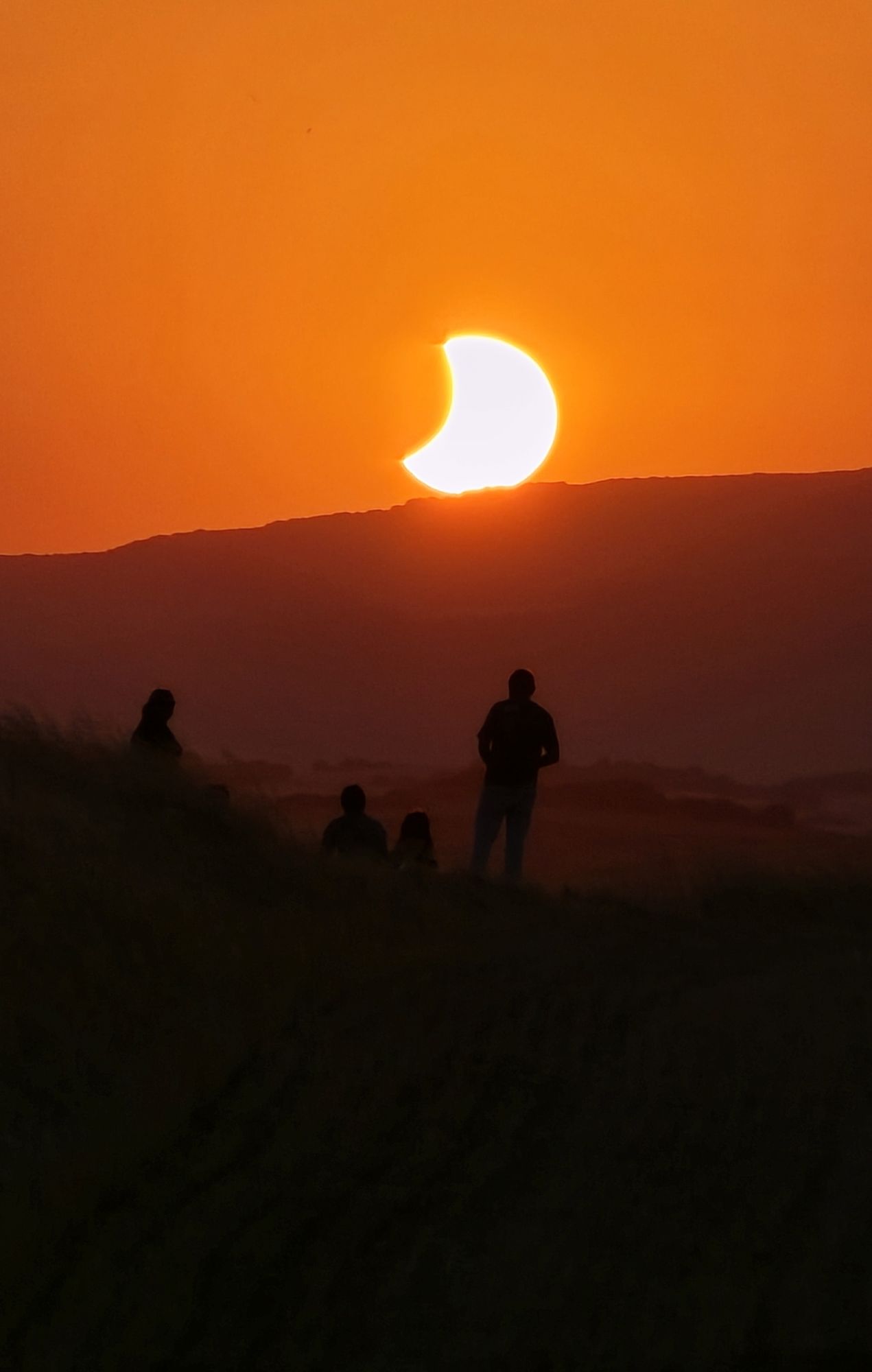 Soleil partiellement éclipsé se couchant derrière les reliefs, avec des observateurs en silhouette