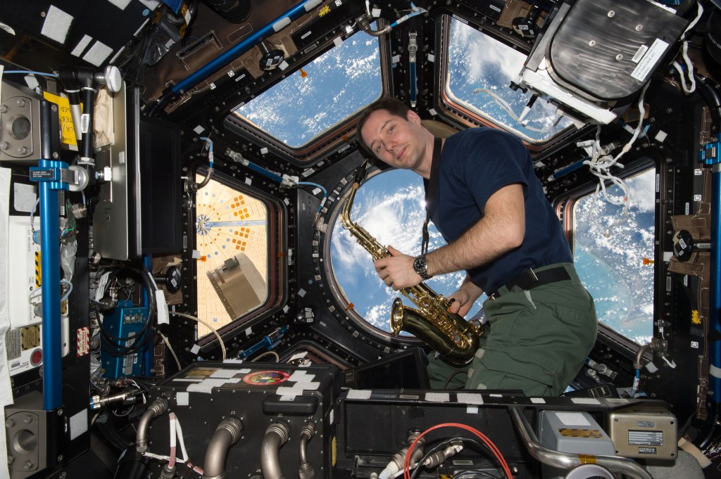 Thomas Pesquet tient un saxophone dans la Coupole de la Station spatiale internationale, avec la Terre visible à travers les hublots.